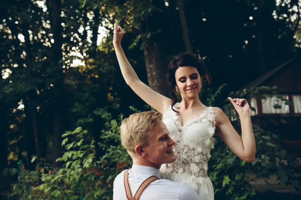 Wedding couple outdoors celebrating joyfully under trees, with bride in lace gown raising arms and partner smiling in suspenders.