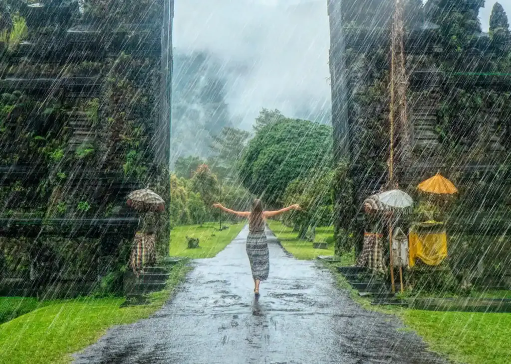 Person standing in rain at Bali temple gates with moss‑covered stone carvings and misty mountains in background